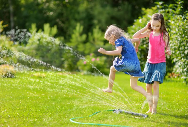 Adorable little girls playing with a sprinkler in a backyard on sunny summer day. Cute children having fun with water outdoors. Funny summer games for kids.